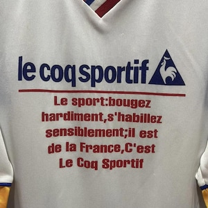 May include: White athletic shirt with a V-neck collar featuring red, white, and blue trim. The shirt displays the text "le coq sportif" in blue, with additional red text below. A blue and white logo is on the right.