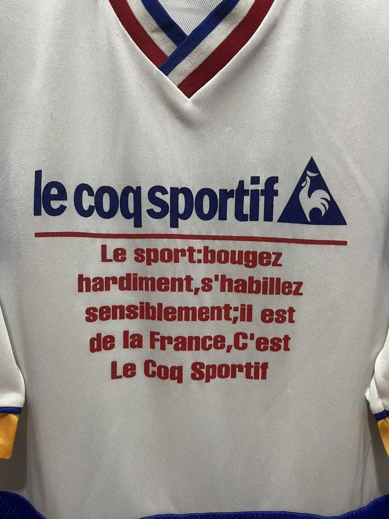 May include: White athletic shirt with a V-neck collar featuring red, white, and blue trim. The shirt displays the text "le coq sportif" in blue, with additional red text below. A blue and white logo is on the right.