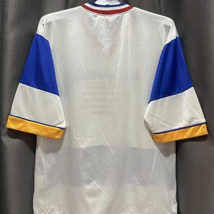 May include: White athletic jersey with blue mesh sleeves and yellow trim. The collar features red, white, and blue stripes. The jersey is hanging on a hanger against a gray background. The back of the jersey is visible.