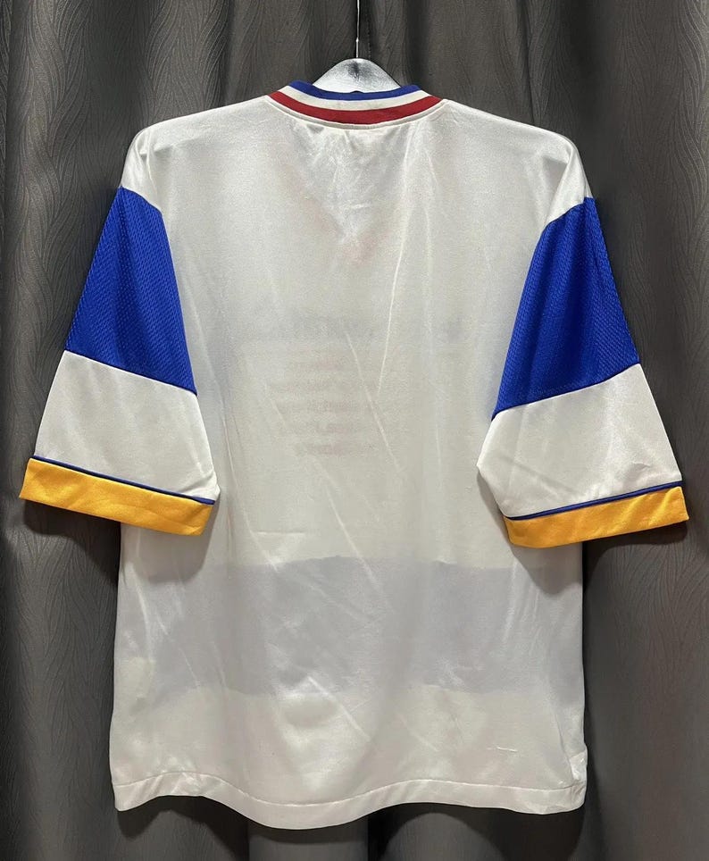 May include: White athletic jersey with blue mesh sleeves and yellow trim. The collar features red, white, and blue stripes. The jersey is hanging on a hanger against a gray background. The back of the jersey is visible.