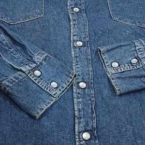 Pode incluir: Close-up de uma camisa de ganga azul. A camisa apresenta dois bolsos no peito, frente abotoada e punhos abotoados. O denim tem um aspeto cl&aacute;ssico e desgastado.