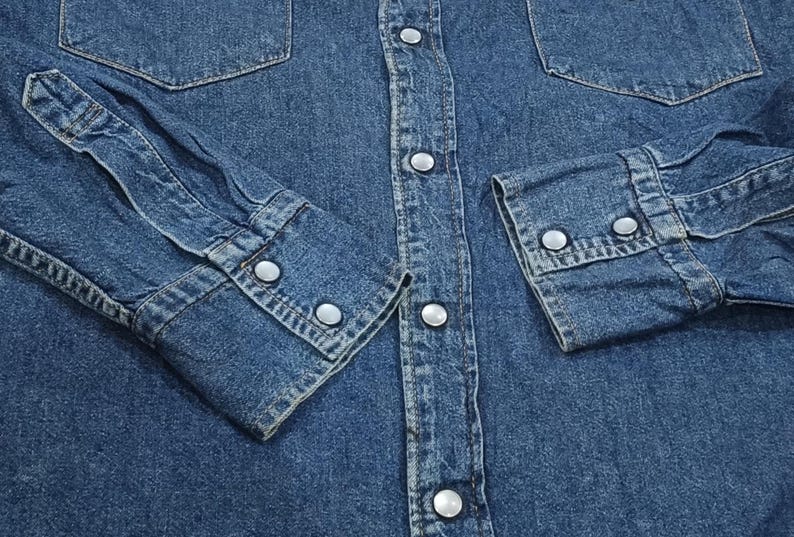 Pode incluir: Close-up de uma camisa de ganga azul. A camisa apresenta dois bolsos no peito, frente abotoada e punhos abotoados. O denim tem um aspeto cl&aacute;ssico e desgastado.