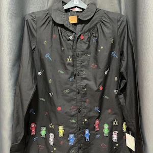 以下が含まれることがあります： 黒いボタンダウンシャツ、ピーターパンカラー、カラフルな刺繍のビデオゲームをテーマにしたデザインが施されています。 シャツには、さまざまなゲームコントローラー、キャラクター、その他のグラフィックが使用されています。 袖口にはタグが付いています。