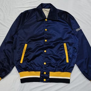 May include: A navy blue satin bomber jacket featuring a pointed collar, yellow snap buttons, and two side pockets with yellow trim. The cuffs and hem are striped in yellow, white, and navy blue. The text OSA is visible on the left sleeve.