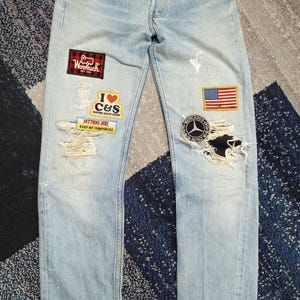 May include: Light blue denim jeans featuring a button fly, distressed knees, and various patches including a red and black plaid Woolrich logo, an I heart C&S emblem, a Setting Sun Keep My Composure patch, a US flag, and a Mercedes-Benz logo.