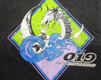 Può includere: T-shirt nera con grafica di drago all'interno di un rombo. Il drago è bianco e nero, su sfondo verde. Sotto, un disegno di onde blu e la scritta "019 SURFBOARDS" in bianco.