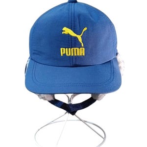 May include: A blue baseball cap with a yellow Puma logo. The cap has a curved brim and ear flaps. The Puma logo is embroidered in yellow thread. The cap is displayed on a white stand.