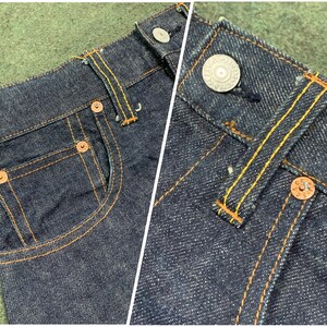 Pu&ograve; includere: Primo piano di jeans in denim blu scuro con rivetti in rame e cuciture gialle. I jeans hanno uno stile classico a cinque tasche con un dettaglio di tasca posteriore.
