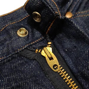 May include: Close-up of the top portion of dark blue denim jeans. Visible details include a brass zipper, metal button, and rivets. The stitching is a contrasting tan color, highlighting the seams and edges of the garment.