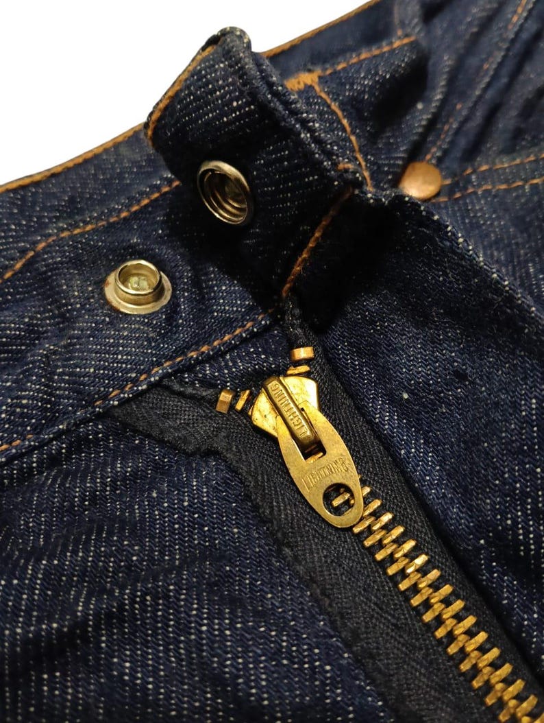 May include: Close-up of the top portion of dark blue denim jeans. Visible details include a brass zipper, metal button, and rivets. The stitching is a contrasting tan color, highlighting the seams and edges of the garment.