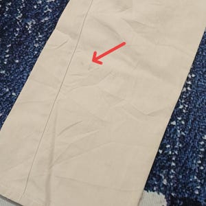 May include: Close-up of beige trousers with a central seam. The fabric is a light, neutral colour, possibly cotton or similar. The trousers are folded, and the fabric has a slightly wrinkled texture.