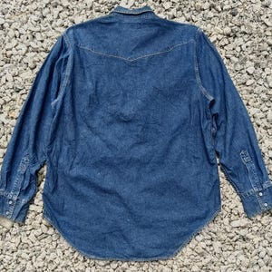 Pode incluir: Camisa de ganga de manga comprida em lavagem azul m&eacute;dio. A camisa apresenta uma gola cl&aacute;ssica, frente abotoada e punhos abotoados. A parte de tr&aacute;s tem um design pontiagudo. A camisa &eacute; feita de tecido de ganga resistente.