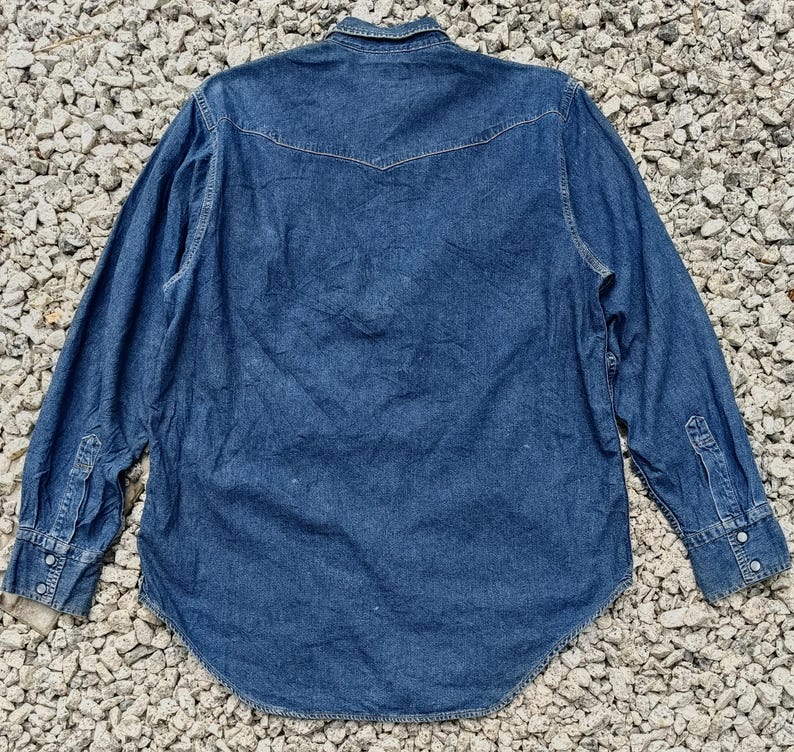 Pode incluir: Camisa de ganga de manga comprida em lavagem azul m&eacute;dio. A camisa apresenta uma gola cl&aacute;ssica, frente abotoada e punhos abotoados. A parte de tr&aacute;s tem um design pontiagudo. A camisa &eacute; feita de tecido de ganga resistente.