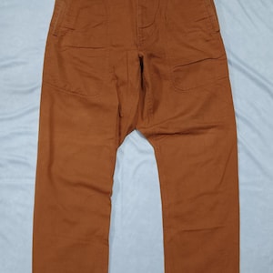May include: A pair of solid rust-colored trousers featuring a high-waisted design with a single dark button closure at the center. These pants include two large, curved front pockets and a straight-leg cut, providing a structured appearance.