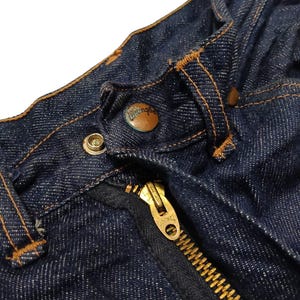 May include: Close-up of a pair of dark blue denim jeans. The image highlights the button, zipper, and stitching details. The zipper is gold-colored, and the button has the word "Wrangler" on it.