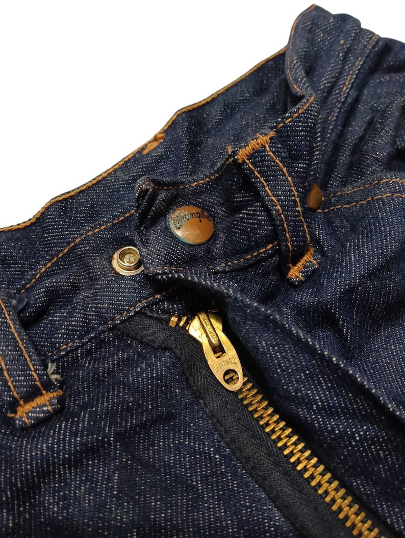 May include: Close-up of a pair of dark blue denim jeans. The image highlights the button, zipper, and stitching details. The zipper is gold-colored, and the button has the word "Wrangler" on it.