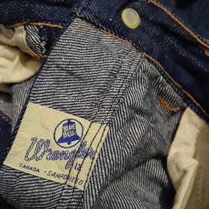 May include: Close-up of blue denim jeans with a visible label. The label reads "Wrangler" with "Blue Bell" and "Canada - Sanforized" text. The jeans have a button and orange stitching.