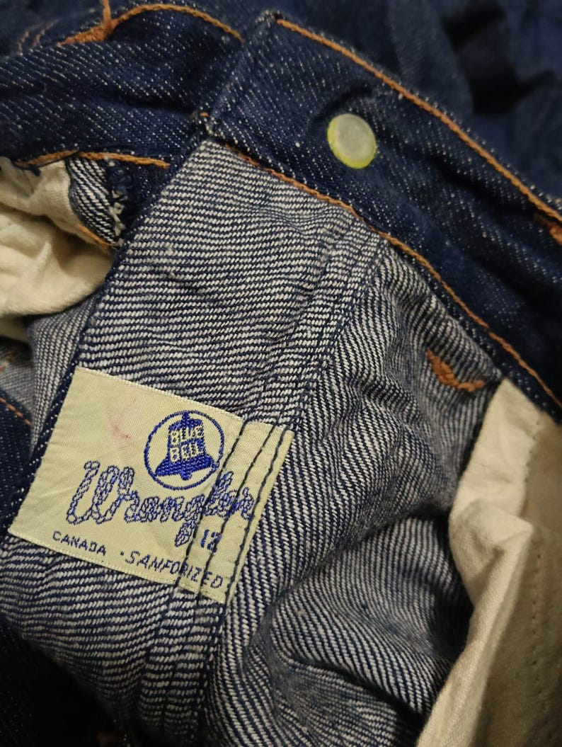 May include: Close-up of blue denim jeans with a visible label. The label reads "Wrangler" with "Blue Bell" and "Canada - Sanforized" text. The jeans have a button and orange stitching.