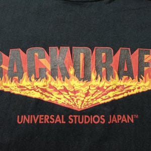Peut inclure: T-shirt noir avec l'inscription "BACKDRAFT" dans un design enflammé en 3D au-dessus de "UNIVERSAL STUDIOS JAPAN" en rouge. Le graphisme comprend des flammes dans les tons de jaune, orange et rouge.