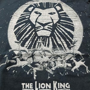 Puede incluir: Camiseta negra con un diseño de El Rey León. El diseño muestra la cara de un león frente a una luna, con gacelas corriendo debajo. Las palabras "THE LION KING" están impresas en blanco en la parte inferior.