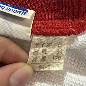 May include: Close-up of a white, red, and blue athletic shirt. The shirt has a red collar and a white label with the text "q sportif". A tag with Japanese text indicates size L, with measurements of 94 and 170.