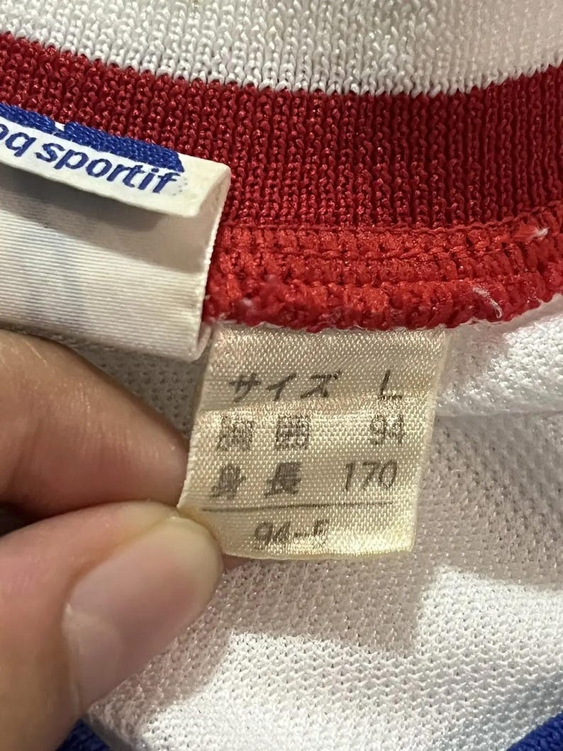 May include: Close-up of a white, red, and blue athletic shirt. The shirt has a red collar and a white label with the text "q sportif". A tag with Japanese text indicates size L, with measurements of 94 and 170.