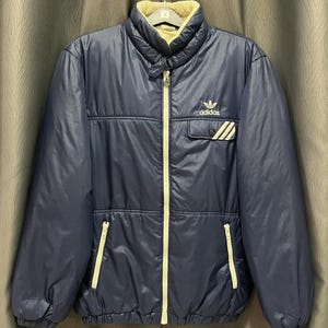 Op de afbeelding: Een marineblauwe Adidas jas met een crèmekleurige kraag en voering. De jas heeft een lange ritssluiting, twee voorzakken en het Adidas-logo op de borst. De jas heeft een gewatteerd ontwerp en een klassieke, sportieve uitstraling.