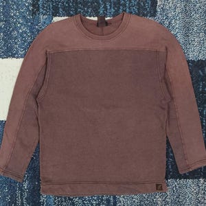 Pode incluir: Uma camisola de cor bord&ocirc; com mangas compridas. A parte superior &eacute; de um tom mais claro de bord&ocirc;, com um painel retangular bord&ocirc; mais escuro no peito. Tem gola redonda e um pequeno log&oacute;tipo no canto inferior direito.