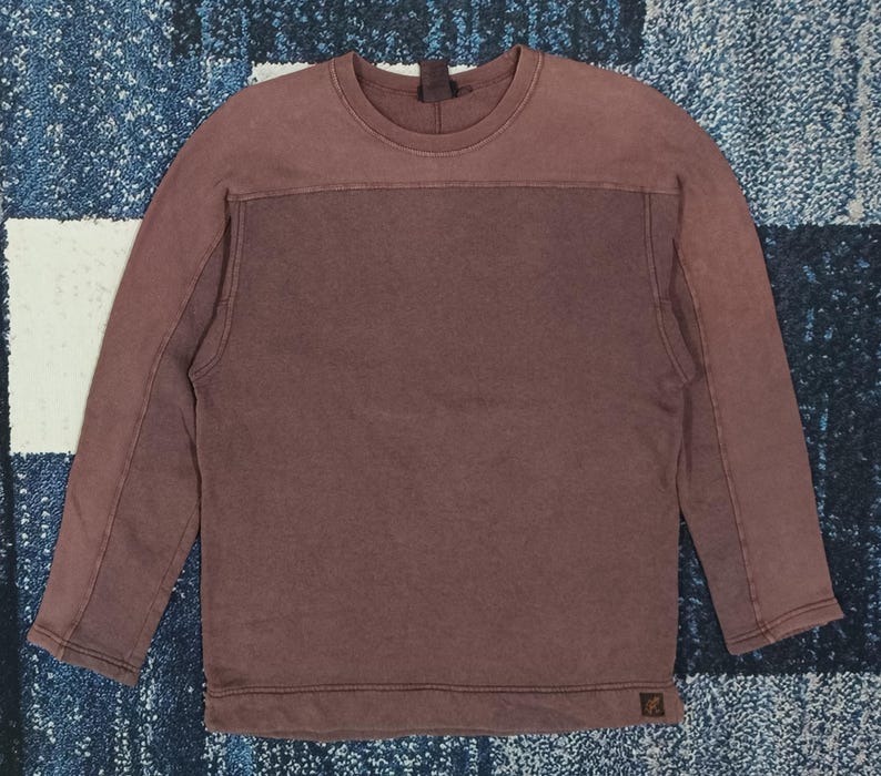 Pode incluir: Uma camisola de cor bord&ocirc; com mangas compridas. A parte superior &eacute; de um tom mais claro de bord&ocirc;, com um painel retangular bord&ocirc; mais escuro no peito. Tem gola redonda e um pequeno log&oacute;tipo no canto inferior direito.