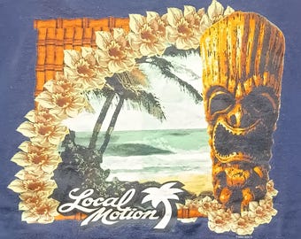 Può includere: T-shirt blu navy con un design tropicale. Presenta una maschera tiki, palme e onde, incorniciate da fiori. Le parole "Local Motion" sono visualizzate in un carattere stilizzato. Il design evoca un tema hawaiano.