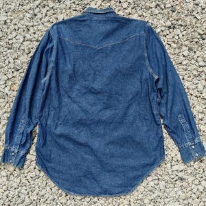 Pode incluir: Camisa de ganga de manga comprida em lavagem azul m&eacute;dia. Apresenta uma frente abotoada, uma gola cl&aacute;ssica e punhos abotoados. A parte de tr&aacute;s tem um design pontiagudo e a bainha &eacute; curva.