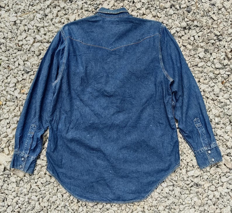Pode incluir: Camisa de ganga de manga comprida em lavagem azul m&eacute;dia. Apresenta uma frente abotoada, uma gola cl&aacute;ssica e punhos abotoados. A parte de tr&aacute;s tem um design pontiagudo e a bainha &eacute; curva.