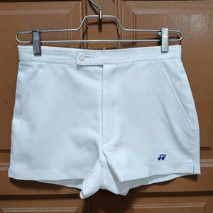 Peut inclure: Shorts de sport blancs avec fermeture boutonnée et poches. Les shorts présentent un petit logo bleu en bas à droite. Ils sont suspendus à un cintre en métal.