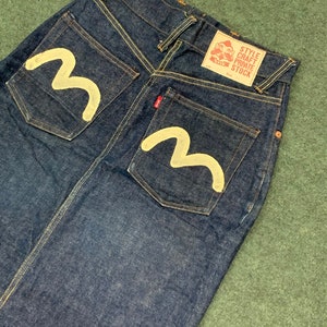 Pu&ograve; includere: Un paio di jeans in denim blu scuro con un design a onda bianca sulle tasche posteriori. I jeans hanno un'etichetta rossa sulla tasca posteriore destra che dice "Style Craft Private Stock".