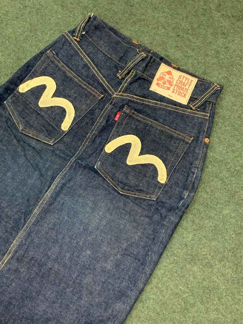 Pu&ograve; includere: Un paio di jeans in denim blu scuro con un design a onda bianca sulle tasche posteriori. I jeans hanno un'etichetta rossa sulla tasca posteriore destra che dice "Style Craft Private Stock".
