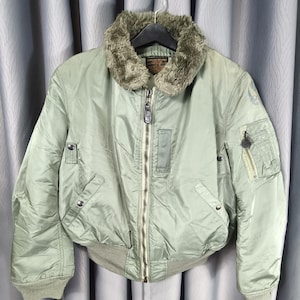 May include: A sage green bomber jacket with a faux fur collar. The jacket features a zip-up front, multiple pockets, and ribbed cuffs and hem. The jacket is hanging on a hanger against a grey curtain.
