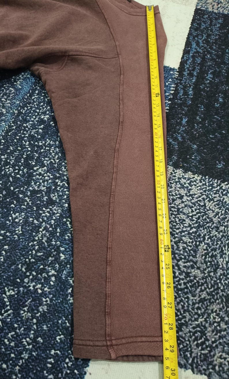 Pode incluir: Uma camisola de cor bord&ocirc; com uma fita m&eacute;trica amarela ao lado. A fita indica medidas em cent&iacute;metros, com o comprimento da pe&ccedil;a a atingir cerca de 76 cm.