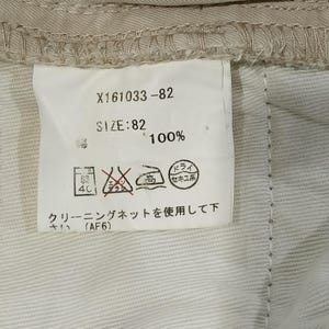 May include: A close-up of a clothing label with the text "X161033-82" and "SIZE: 82". The label also includes washing instructions and the text "100%". The label is attached to beige fabric.