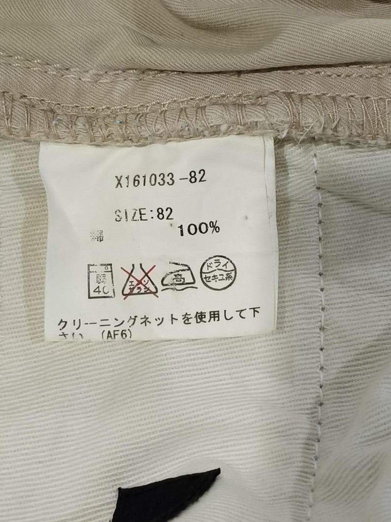 May include: A close-up of a clothing label with the text "X161033-82" and "SIZE: 82". The label also includes washing instructions and the text "100%". The label is attached to beige fabric.