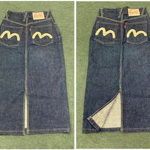Pu&ograve; includere: Una gonna in denim blu scuro con uno spacco sul davanti. La gonna ha due tasche posteriori con un logo ricamato bianco che dice "RELAX" e una forma stilizzata a "M".
