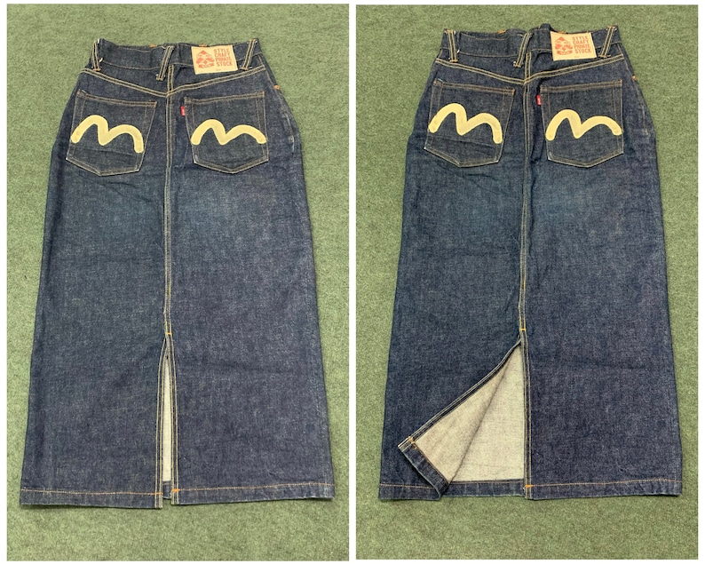Pu&ograve; includere: Una gonna in denim blu scuro con uno spacco sul davanti. La gonna ha due tasche posteriori con un logo ricamato bianco che dice "RELAX" e una forma stilizzata a "M".