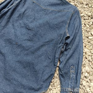 Pode incluir: Camisa de ganga azul desbotada com mangas compridas. Apresenta gola cl&aacute;ssica, punhos abotoados e detalhe de pala nas costas. O tecido mostra sinais de desgaste, conferindo-lhe um aspeto vintage.