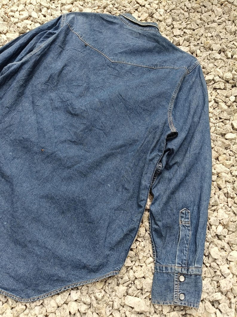 Pode incluir: Camisa de ganga azul desbotada com mangas compridas. Apresenta gola cl&aacute;ssica, punhos abotoados e detalhe de pala nas costas. O tecido mostra sinais de desgaste, conferindo-lhe um aspeto vintage.