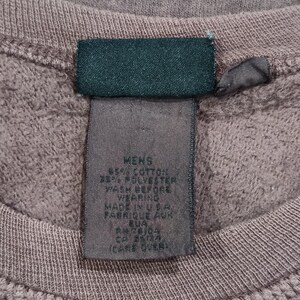 Pode incluir: Close-up de um moletom malva com bainha canelada e etiqueta verde escuro. A etiqueta diz "MENS" e lista a composi&ccedil;&atilde;o do tecido e instru&ccedil;&otilde;es de cuidado. Feito nos EUA.