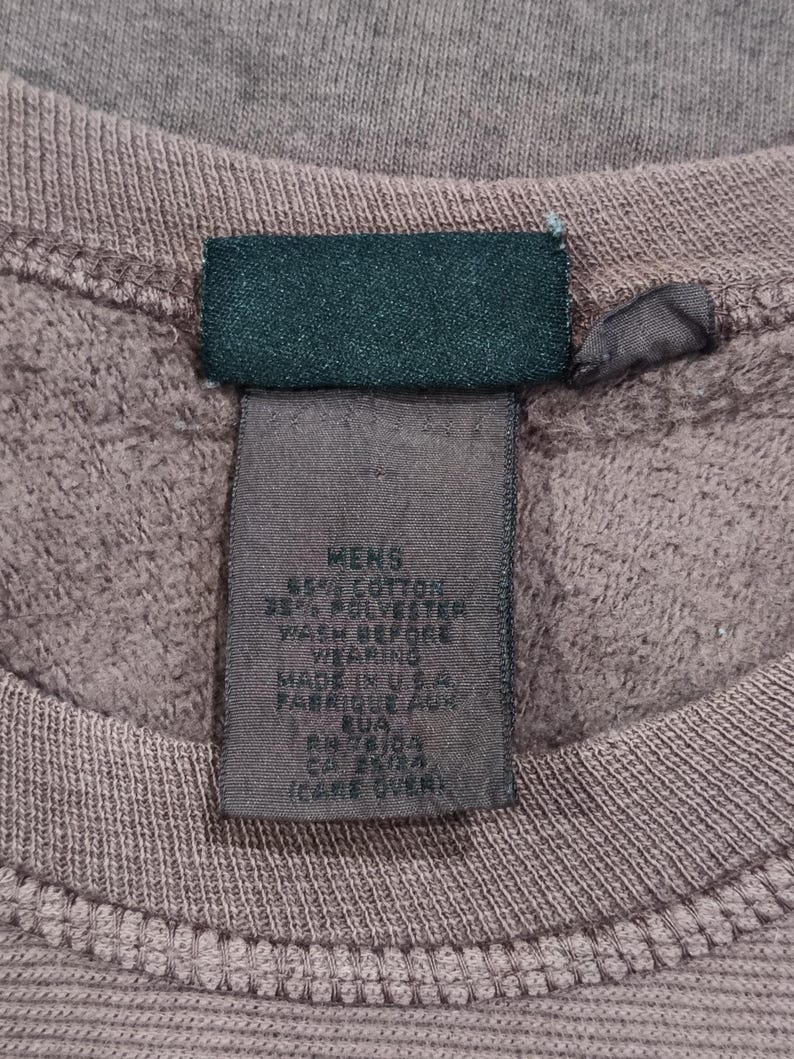 Pode incluir: Close-up de um moletom malva com bainha canelada e etiqueta verde escuro. A etiqueta diz "MENS" e lista a composi&ccedil;&atilde;o do tecido e instru&ccedil;&otilde;es de cuidado. Feito nos EUA.