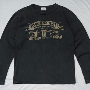 May include: A black long-sleeve crew neck shirt featuring a tan graphic print on the chest. The design includes the text STUDIO D'ARTISAN, DESIGEN ONLY ORIGINAL DOUBLE, and PIG MARK, along with illustrations of two pigs and a pair of jeans.