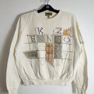 May include: Cream-coloured sweatshirt with the text "KENZO GOLF" in a grid pattern. The design features various colours and textures within the letters and grid. The sweatshirt has a crew neck and long sleeves.