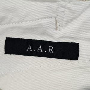 May include: Close-up of off-white fabric with visible stitching. A black rectangular label is sewn onto the fabric, displaying the letters "A.A.R" in white. The fabric appears to be part of a garment.