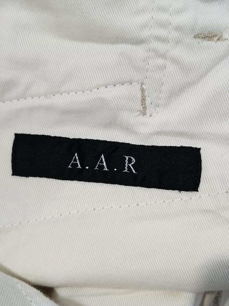 May include: Close-up of off-white fabric with visible stitching. A black rectangular label is sewn onto the fabric, displaying the letters "A.A.R" in white. The fabric appears to be part of a garment.