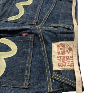 Pu&ograve; includere: Jeans in denim blu con un designo ricamato bianco sulle tasche posteriori. I jeans hanno una toppa in pelle marrone con il testo "Style Craft Private Stock EVIS" e "Lot. 010 Taglia 36".